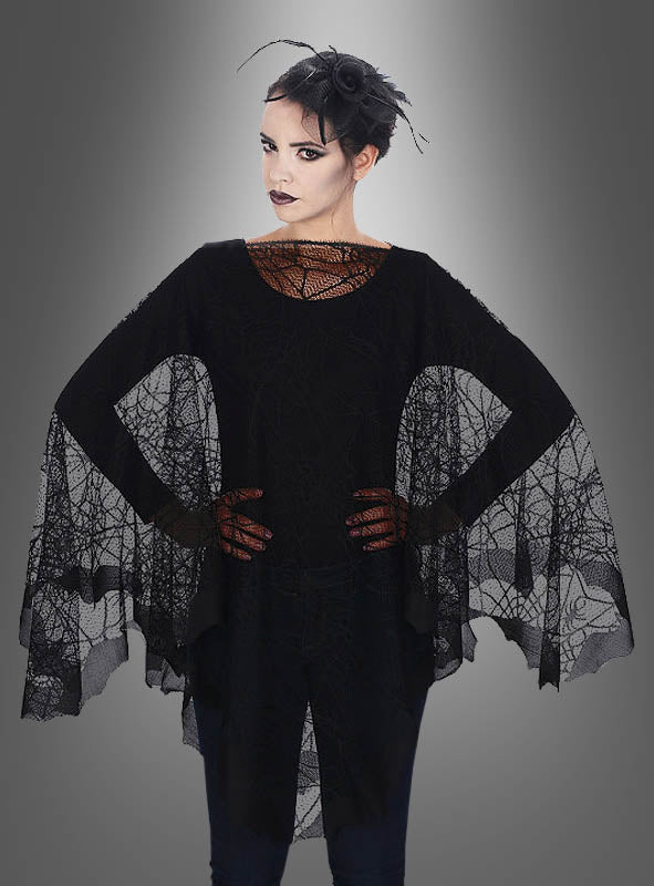 Eine Person, die den „Filigraner Spinnennetz-Poncho“ trägt – mit schwarzem Spinnennetzmuster, ausgestellten Ärmeln, dunklem Make-up und einem Kopfschmuck aus Federn –, steht vor einem grauen Hintergrund mit Farbverlauf.