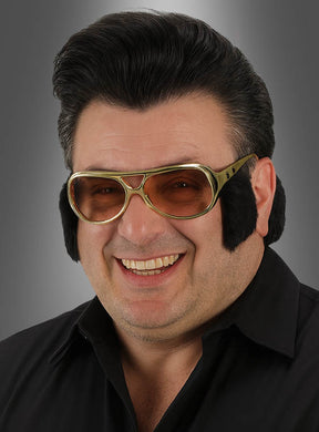 Ein Mann mit dunklem Haar trägt die goldfarbene 70er-Brille mit Koteletten – eine große goldene Sonnenbrille mit schwarzen Koteletten – sowie ein schwarzes Hemd und lächelt vor einem grauen Hintergrund mit Farbverlauf.