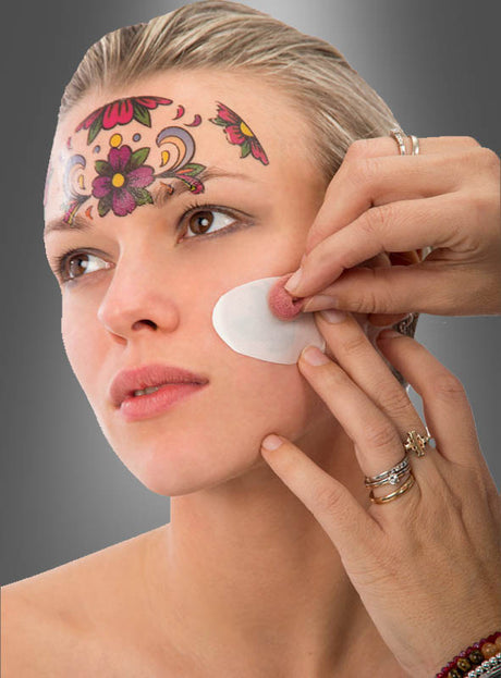 Eine Person trägt mit einem Wattepad Make-up auf die Wange einer Frau auf. Die Frau trägt „Gesichts-Tattoos zum Tag der Toten“ – farbenfrohe Blumenmotive auf der Stirn, die von den leuchtenden Sugar-Skull-Motiven inspiriert sind, wie man sie oft bei den Feierlichkeiten zum Tag der Toten sieht.