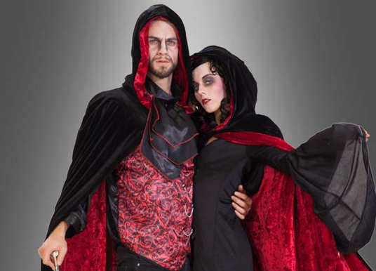 Zwei Personen, die einheitliche rot-schwarze Vampir-Kostüme mit Kapuzenumhängen tragen, posieren gemeinsam vor einem grauen Hintergrund.