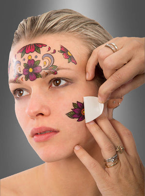 Eine Person bringt ein temporäres Blumentattoo auf ihrem Gesicht an, das die bereits vorhandenen „Gesichts-Tattoos zum Tag der Toten“ mit Sugar-Skull-Motiven ergänzt, die ihre Stirn und Wange zieren.