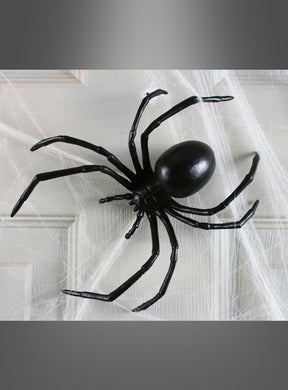 Eine übergroße Schwarze-Witwe-Spinne (15 cm) aus schwarzem Kunststoff liegt auf weißem Spinnennetz-Material vor einer weißen Tür und ist die perfekte Halloween-Dekoration.