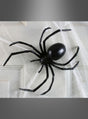 Eine übergroße Schwarze-Witwe-Spinne (15 cm) aus schwarzem Kunststoff liegt auf weißem Spinnennetz-Material vor einer weißen Tür und ist die perfekte Halloween-Dekoration.
