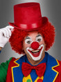 Eine als Clown verkleidete Person mit einer roten Lockenperücke, einer roten Nase, einem bunten Anzug und weißen Handschuhen zieht ihren roten Clown-Zylinderhut leicht zur Seite und lächelt breit vor einem grauen Hintergrund.