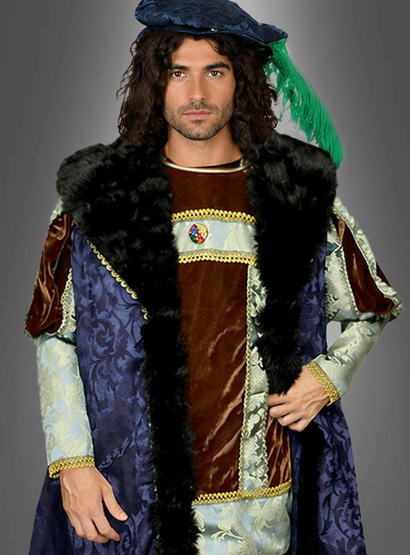 Ein Mann, gekleidet im „Deluxe Großherzog Herrenkostüm“, posiert vor einem schlichten grauen Hintergrund. Er trägt ein von der Renaissance inspiriertes Outfit, bestehend aus einem blauen Umhang, einer braunen Samt-Tunika mit Pelzbesatz und einem blauen Hut, der mit einer grünen Feder verziert ist. Dieses elegante Kostüm verkörpert authentisch den Stil des Adels der Renaissance.