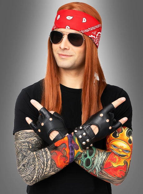 Eine Person mit langen roten Haaren, einem roten Bandana, Sonnenbrille, schwarzen Handschuhen und Tattoo-Ärmeln verschränkt die Arme und zeigt das Rock-Handzeichen vor grauem Hintergrund – perfekt für alle, die das 7-teilige Rocker-Kostüm-Set suchen oder ein authentisches 80er-Jahre-Outfit lieben.