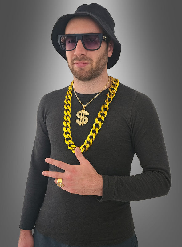 Ein Mann mit schwarzem Hut, Sonnenbrille, schwarzem Hemd und großen Goldketten – darunter eine Halskette mit Dollarzeichen aus dem „Pimp Rapper 80er Set“ (5-teilig) – steht vor einem grauen Hintergrund mit Farbverlauf und formt mit den Fingern ein Peace-Zeichen.