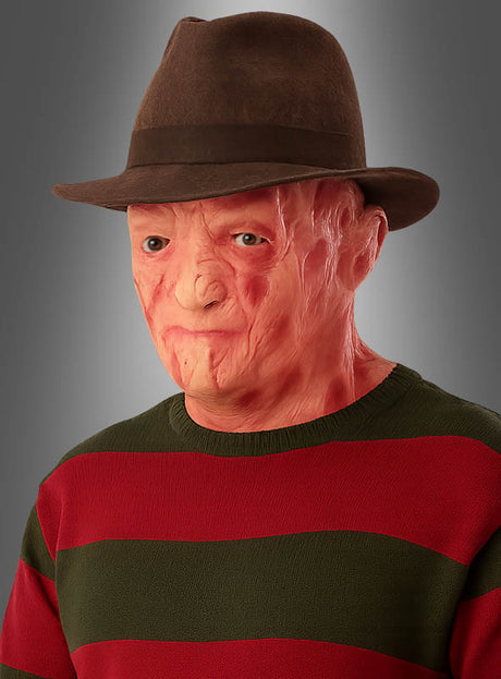 Eine Person, die einen braunen Fedora-Hut, einen grün-rot gestreiften Pullover und die Maske „Klingenmörder Freddy“ trug – die einem verbrannten Gesicht ähnelt und von einer Horrorfilm-Maske inspiriert ist –, posierte vor einem grauen Hintergrund.