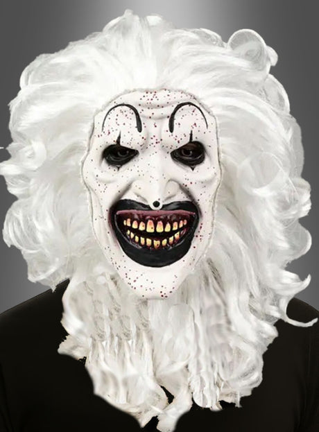 Eine Person, die die schwarz-weiße Clownmaske mit Haaren trägt – mit einem grotesken schwarz-weißen Design, einem übertrieben großen schwarzen Mund, gelben Zähnen, geschwungenen Augenbrauen, roten Flecken und langen weißen Locken –, steht vor einem grauen Hintergrund.