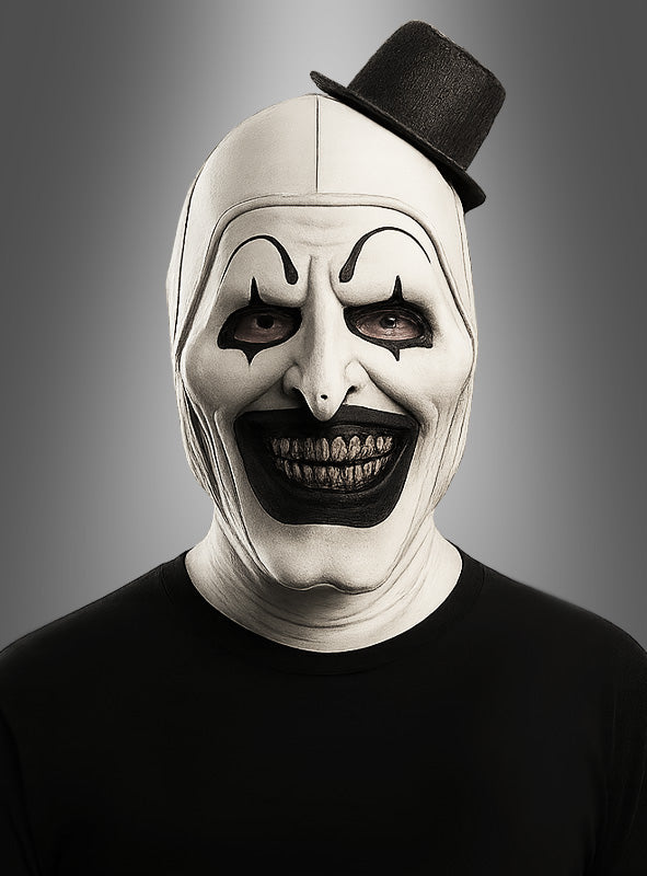 Eine Person, die eine schwarz-weiße Clownmaske mit Zylinder trägt, mit übertriebenem schwarzem Make-up, einem breiten Grinsen und einem kleinen, schräg sitzenden schwarzen Hut, vor einem grauen Hintergrund.
