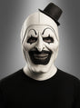 Eine Person, die eine schwarz-weiße Clownmaske mit Zylinder trägt, mit übertriebenem schwarzem Make-up, einem breiten Grinsen und einem kleinen, schräg sitzenden schwarzen Hut, vor einem grauen Hintergrund.