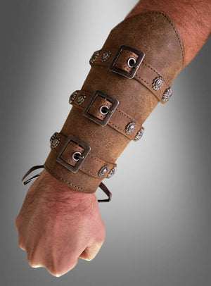 Vor einem grauen Hintergrund mit Farbverlauf ist ein Unterarm zu sehen, an dem ein brauner Armschutz im mittelalterlichen Antik-Look mit vier Metallschnallen und Ziernieten getragen wird. Dieses Accessoire für Ritterkostüme verleiht jedem Outfit im historischen Stil einen authentischen mittelalterlichen Touch.