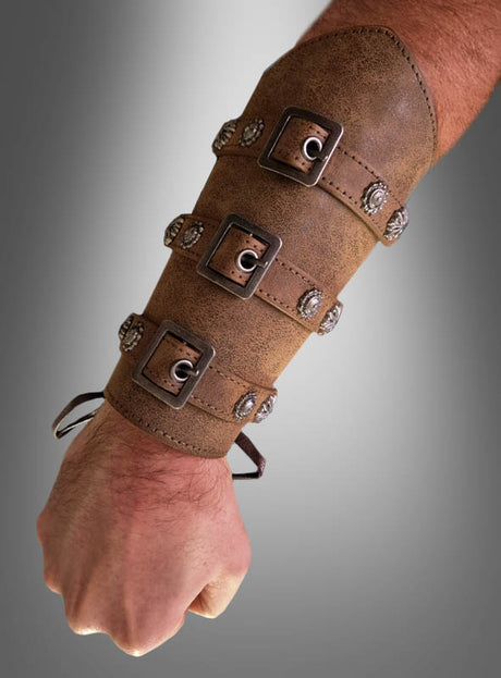 Vor einem grauen Hintergrund mit Farbverlauf ist ein Unterarm zu sehen, an dem ein brauner Armschutz im mittelalterlichen Antik-Look mit vier Metallschnallen und Ziernieten getragen wird. Dieses Accessoire für Ritterkostüme verleiht jedem Outfit im historischen Stil einen authentischen mittelalterlichen Touch.