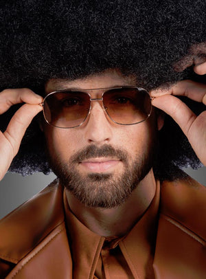 Ein Mann mit einer voluminösen Afro-Frisur, der eine braune Jacke und eine stylische braune Retro-Sonnenbrille im 70er-Jahre-Stil trägt, blickt direkt in die Kamera, während er seine Brille zurechtrückt.