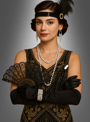Diese Frau trägt ein Flapper-Outfit aus den 1920er Jahren und ergänzt ihren Look mit schwarzen Handschuhen, einem perlenbesetzten Stirnband, Perlenschmuck und einem Spitzenfächer – ihr elegantes Gatsby-Kostüm wird durch das 20er-Jahre-Accessoire-Set in Schwarz mit Fächer (9-teilig) perfekt abgerundet –, während sie vor einem grauen Hintergrund steht.