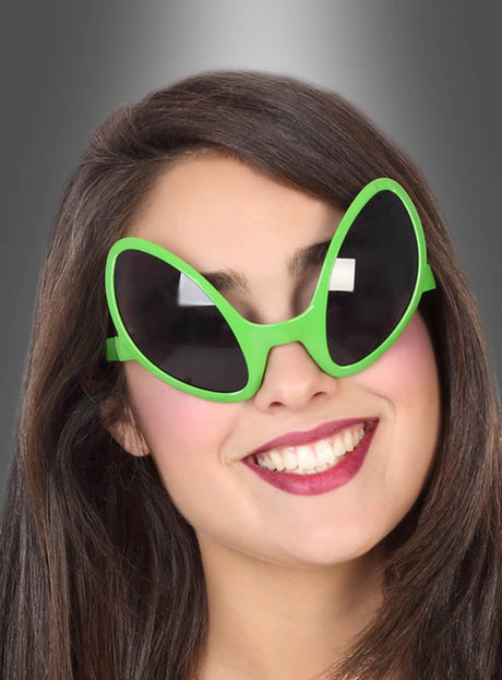 Eine Frau lächelt, während sie die „Alien Brille“ in Grün mit getönten Gläsern trägt, deren große, grüne, ovale Gläser an die Augen eines Außerirdischen erinnern und sich vor einem grau verlaufenden Hintergrund abheben.