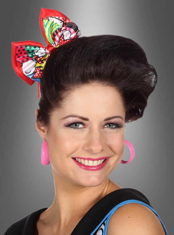 Eine lächelnde Frau mit gestyltem dunklem Haar und großen rosa Creolen trägt die farbenfrohe Pop-Art-Haarschleife als verspieltes Haaraccessoire und posiert vor einem grauen Hintergrund mit Farbverlauf.