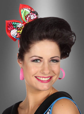 Eine lächelnde Frau mit gestyltem dunklem Haar und großen rosa Creolen trägt die farbenfrohe Pop-Art-Haarschleife als verspieltes Haaraccessoire und posiert vor einem grauen Hintergrund mit Farbverlauf.