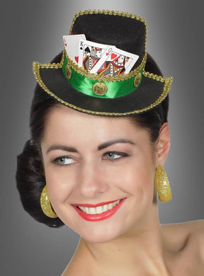 Eine Frau, die den „Casino Mini Hut am Haarreif“ trägt – einen schwarz-grün gesäumten Miniaturhut, verziert mit Spielkarten und goldenen Akzenten, dazu passende goldene Ohrringe –, lächelt vor einem grauen Hintergrund und macht ihn damit zur perfekten Ergänzung für jedes Las-Vegas-Kostüm oder jede Damen-Accessoire-Kollektion.