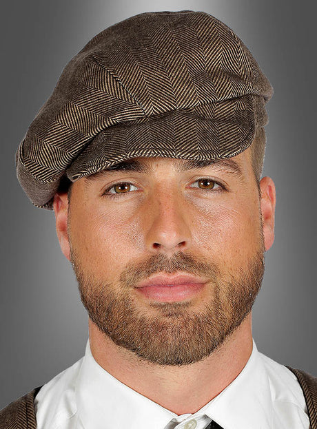 Ein Mann mit gepflegtem Bart trägt die „Braune Schiebermütze im Peaky-Blinders-Stil“ im Fischgrätenmuster sowie ein weißes Hemd und eine braune Weste und blickt vor einem grauen Hintergrund nach vorne – für einen authentischen Peaky-Blinders-Look.