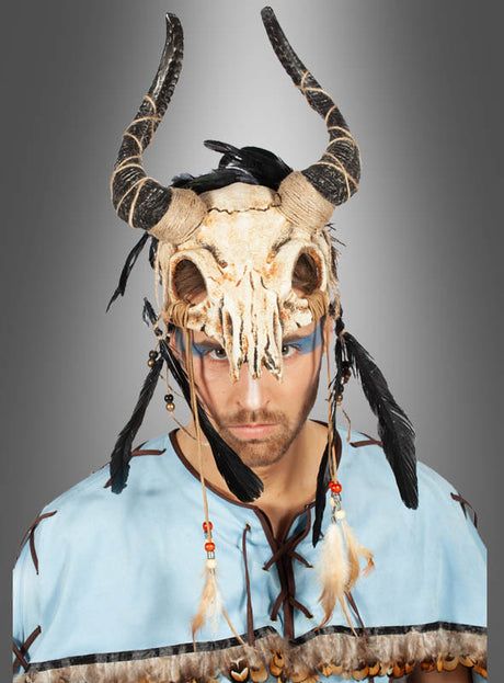 Eine Person, die eine mit Federn und Perlen verzierte „Stier-Skelett-Maske mit Hörnern“ trägt und ein hellblaues Oberteil mit Lederschnürung anhat, blickt vor einem grauen Hintergrund mit Farbverlauf geradeaus.
