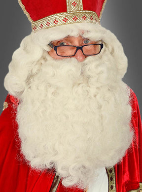 Eine Person, die eine rot-goldene Robe und eine Mitra trägt, mit der „Deluxe Weihnachtsmann-Perücke und Bart“ – einem Set aus einer großen, weißen, lockigen Perücke und einem Bart – sowie einer Brille, blickt vor einem grauen Hintergrund in die Kamera.
