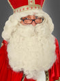 Eine Person, die eine rot-goldene Robe und eine Mitra trägt, mit der „Deluxe Weihnachtsmann-Perücke und Bart“ – einem Set aus einer großen, weißen, lockigen Perücke und einem Bart – sowie einer Brille, blickt vor einem grauen Hintergrund in die Kamera.