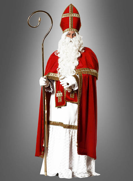 Eine Person, die das Luxus-Nikolaus-Kostüm aus Plüsch trägt – bestehend aus einem roten Umhang, einem weißen Bart, einer Bischofsmütze und einem goldenen Stab –, steht vor einem schlichten grauen Hintergrund.