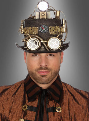 Ein Mann, der eine braune Jacke und den „Steampunk-Zylinder mit Grubenlampe“ trägt – einen äußerst detailreichen Hut mit Zahnrädern, Anzeigen, Spulen und metallischen Akzenten, der von alten Grubenlampen inspiriert ist –, steht vor einem grauen Hintergrund.