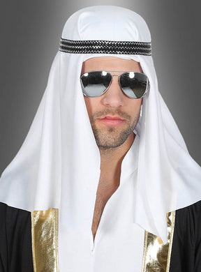 Ein Mann, der die Scheich-Kopfbedeckung im Orient mit einem weißen Schleier, einem schwarz-goldenen Gewand und einer Pilotenbrille trägt, steht vor einem grauen Hintergrund und vermittelt die Eleganz eines Orient-Kostüms.