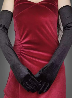 Eine Person in einem roten Samtkleid mit gerafften Seitendetails, die lange, schwarze Samthandschuhe aus elegantem schwarzem Satin trägt und vor einem schlichten grauen Hintergrund steht.