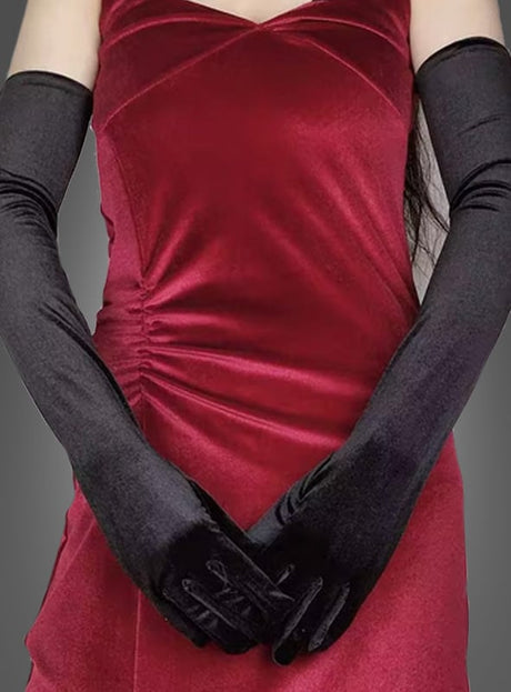 Eine Person in einem roten Samtkleid mit gerafften Seitendetails, die lange, schwarze Samthandschuhe aus elegantem schwarzem Satin trägt und vor einem schlichten grauen Hintergrund steht.