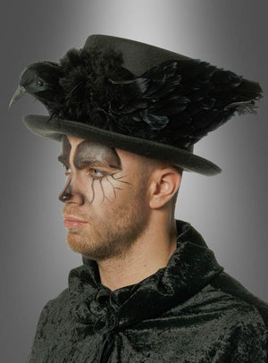 Ein Mann trägt einen schwarzen Samtumhang und den „Zylinder mit Rabe Gothic“ – einen schwarzen Zylinder, der mit Federn, einer Rabenverzierung und Steampunk-Kostümaccessoires geschmückt ist – sowie schwarze Gesichtsbemalung, die einer Vogelmaske ähnelt.