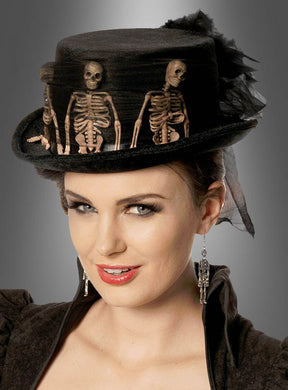 Eine Frau trägt den „Gothic Samt Zylinder mit Skeletten Deluxe“, der als Halloween-Zylinder für Damen gestaltet und mit kleinen Skelettfiguren sowie passenden Skelettohrringen verziert ist, vor einem neutralen grauen Hintergrund – das perfekte Accessoire für alle, die die Ästhetik des Gothic-Huts lieben.