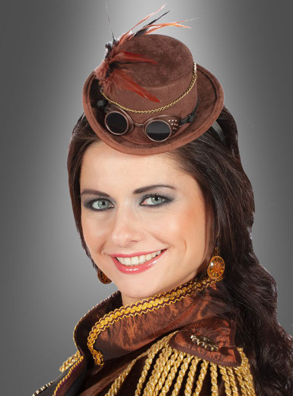 Eine Frau mit langen, dunklen Haaren trägt den „Steampunk Mini Zylinder mit Brille“, der mit einer Schutzbrille und Federn verziert ist, sowie kunstvolle Ohrringe und eine aufwendig gearbeitete braun-goldene Jacke; sie lächelt vor einem grauen Hintergrund mit Farbverlauf.