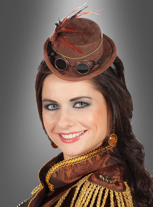 Eine Frau mit langen, dunklen Haaren trägt den „Steampunk Mini Zylinder mit Brille“, der mit einer Schutzbrille und Federn verziert ist, sowie kunstvolle Ohrringe und eine aufwendig gearbeitete braun-goldene Jacke; sie lächelt vor einem grauen Hintergrund mit Farbverlauf.