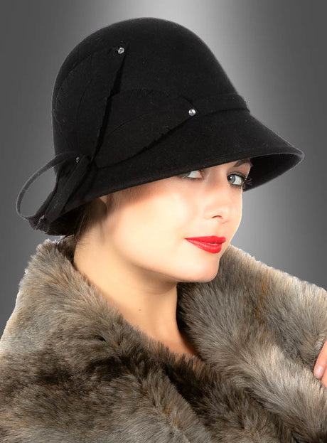 Eine Frau posiert im Profil vor einem grauen Hintergrund mit Farbverlauf; sie trägt einen Pelzmantel und den Cloche-Hut „Luxus Filzhut 20er Jahre schwarz“.
