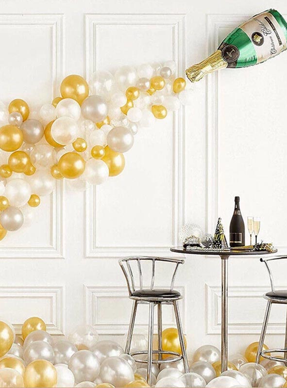 Goldene, weiße und silberne Luftballons („Ballons gold silber“) ergießen sich aus der Wanddekoration „Partyset Champagnerflasche Sekt Deko 42 Teile“ und schaffen so eine beeindruckende Partykulisse mit einem kleinen runden Tisch und zwei Stühlen, die davor stehen.