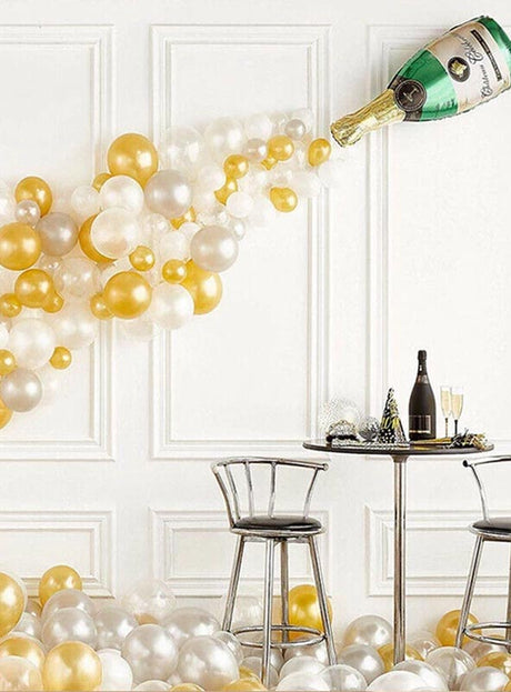 Goldene, weiße und silberne Luftballons („Ballons gold silber“) ergießen sich aus der Wanddekoration „Partyset Champagnerflasche Sekt Deko 42 Teile“ und schaffen so eine beeindruckende Partykulisse mit einem kleinen runden Tisch und zwei Stühlen, die davor stehen.