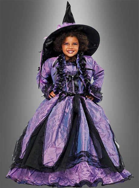 Ein Kind trägt ein lila-schwarzes „Kleine Hexe Sofia“-Kinderkostüm und steht lächelnd vor einem grauen Hintergrund mit Farbverlauf.