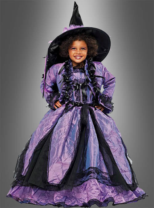 Ein Kind trägt ein lila-schwarzes „Kleine Hexe Sofia“-Kinderkostüm und steht lächelnd vor einem grauen Hintergrund mit Farbverlauf.