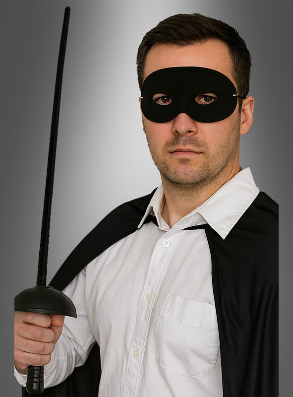 Ein Mann, der das „Bandit Set Maske und Degen 67 cm“ trägt – komplett mit schwarzer Augenmaske und Umhang –, hält ein Schwert aufrecht in der Hand, während er vor einem grauen Hintergrund ein weißes Hemd trägt – was an den ikonischen Stil klassischer Zorro-Kostümaccessoires erinnert.