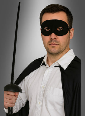Ein Mann, der das „Bandit Set Maske und Degen 67 cm“ trägt – komplett mit schwarzer Augenmaske und Umhang –, hält ein Schwert aufrecht in der Hand, während er vor einem grauen Hintergrund ein weißes Hemd trägt – was an den ikonischen Stil klassischer Zorro-Kostümaccessoires erinnert.