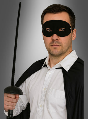 Ein Mann, der das „Bandit Set Maske und Degen 67 cm“ trägt – komplett mit schwarzer Augenmaske und Umhang –, hält ein Schwert aufrecht in der Hand, während er vor einem grauen Hintergrund ein weißes Hemd trägt – was an den ikonischen Stil klassischer Zorro-Kostümaccessoires erinnert.