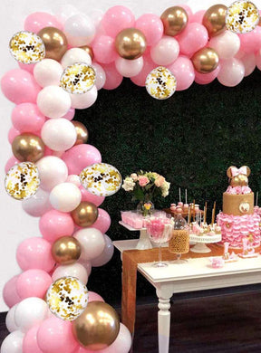 Ein Partytisch mit Desserts und einem „Ballonkette“-Dekoset aus 115 Teilen in Rosa, Gold und Weiß, das einen festlichen Ballonbogen als Hintergrund bildet; außerdem stehen Blumendekorationen und eine Torte auf dem Tisch.