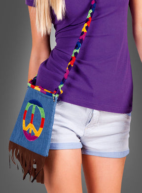 Eine Person, die ein lila Shirt und helle Jeansshorts trägt, hat die „Hippie Jeans Handtasche“ dabei – eine kleine Jeanshandtasche mit Fransen, einem Regenbogen-Friedenssymbol und einem bunten, geflochtenen Riemen, die sich perfekt als Accessoire für ein Hippie-Kostüm eignet.