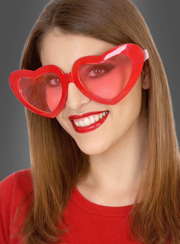 Eine Frau mit glattem braunem Haar trägt die „Riesige Herzbrille XXL“ in leuchtendem Rot zusammen mit einem passenden Oberteil und lächelt vor einem grauen Hintergrund in die Kamera – die perfekte Partybrille und das ideale Karnevalsaccessoire, um jeden festlichen Look abzurunden.