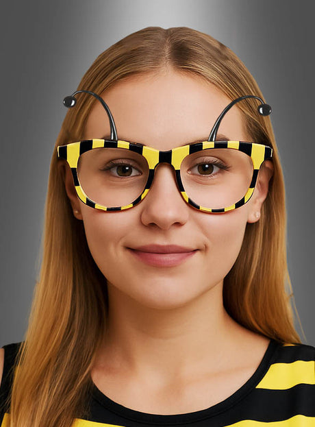 Eine Frau, die die „Brille für Biene“ in Gelb-Schwarz trägt – eine ausgefallene Brille mit gelb-schwarzen Streifen und angebrachten Fühlern –, kombiniert diese mit ihrem gestreiften Shirt und schafft so vor einem grauen Hintergrund das perfekte Accessoire für ihr Bienenkostüm.