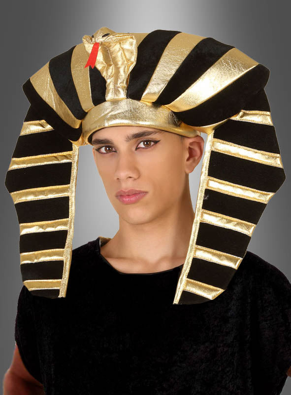 Eine Person, die einen schwarz-goldenen ägyptischen Kopfschmuck trägt, blickt vor einem grauen Hintergrund mit Farbverlauf in die Kamera.