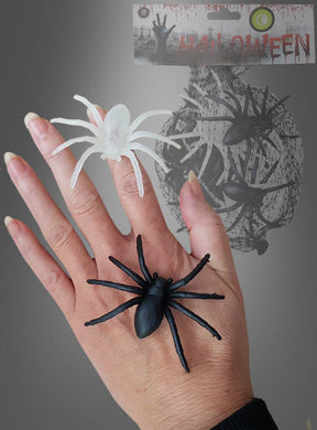 Auf einer Hand mit langen Nägeln sitzen eine schwarze und eine weiße Spinne aus dem 20er-Pack „Spinnen 6 cm – Halloween-Deko“, während im Hintergrund weitere Halloween-Spinnen und themenbezogene Verpackungen zu sehen sind – so entsteht die perfekte gruselige Halloween-Dekoration.